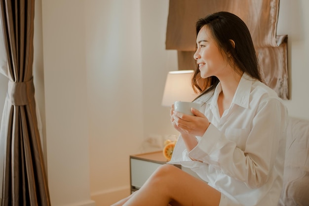 Giovane donna asiatica che vive a casa rilassandosi e bevendo una tazza di caffè caldo in camera da letto in vacanza. Asiatico, asia, relax, da solo, tecnologia, concetto di stile di vita.