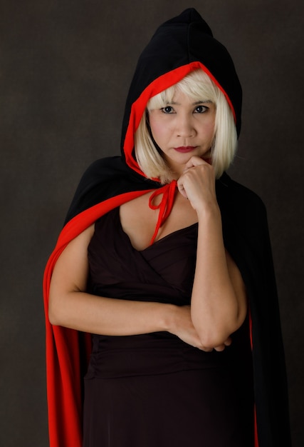 Giovane donna asiatica con capelli biondi che indossa un mantello nero e rosso con cappuccio durante la mascherata di Halloween che guarda la telecamera