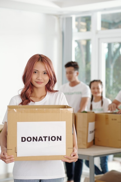 Giovane donna asiatica con i capelli rosa che tiene in mano una grande scatola di generi alimentari e vestiti che le persone hanno donato alla fondazione di beneficenza