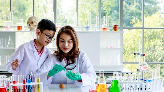 Giovane donna asiatica con lente d'ingrandimento che esamina l'uovo di gallina mentre il ragazzo concentrato mescola liquidi colorati durante l'esperimento scientifico nel laboratorio di chimica.