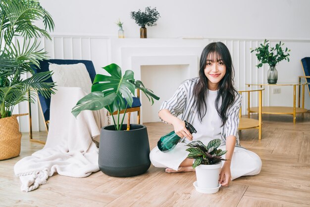 Giovane donna asiatica con lunghi capelli neri che indossa un panno casual spruzzato acqua su una piccola pianta da casa nel vaso con cura Monstera e amante delle piante da casa a casa Il concetto di cura delle piante