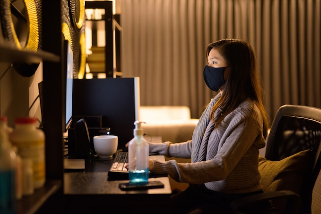 Giovane donna asiatica con maschera che lavora da casa a tarda notte