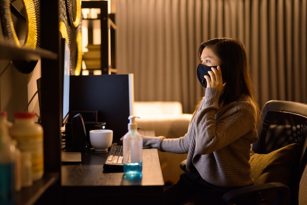 Giovane donna asiatica con maschera parlando al telefono mentre si lavora da casa durante la notte