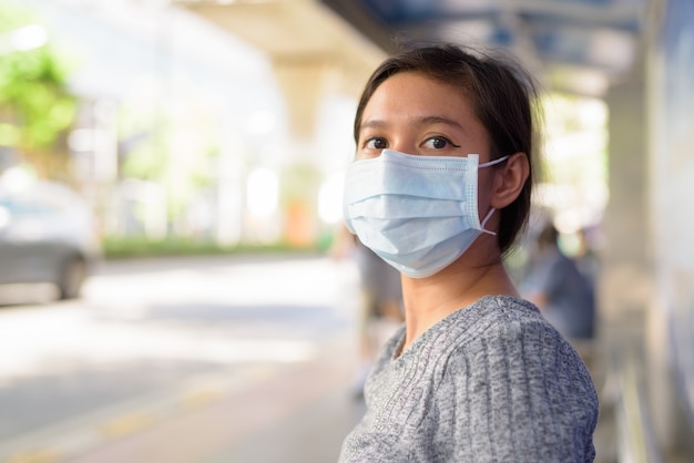 Giovane donna asiatica con maschera per la protezione dallo scoppio del virus corona seduto alla fermata dell'autobus