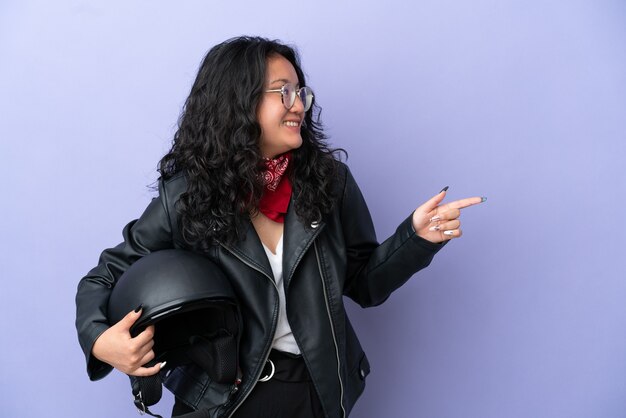 Giovane donna asiatica con un casco da motociclista isolato su sfondo viola che punta il dito di lato e presenta un prodotto