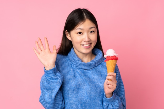 Giovane donna asiatica con un gelato della cornetta sulla parete rosa che saluta con la mano con l'espressione felice