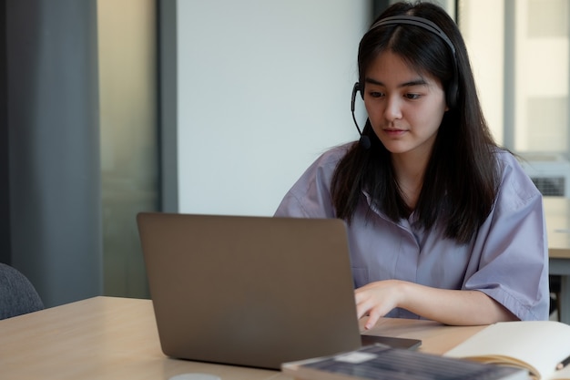 Giovane donna asiatica concentrata con la cuffia avricolare che esamina laptop