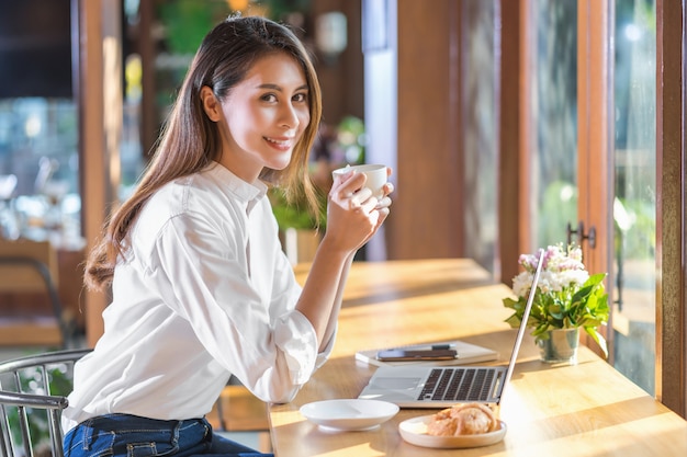 Giovane donna asiatica del ritratto che tiene e che beve una tazza di caffè e che lavora con il computer portatile di tecnologia ad una caffetteria