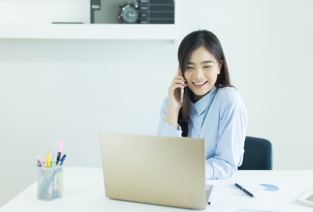 Giovane donna asiatica di affari che parla sul telefono cellulare e che sorride al suo posto di lavoro