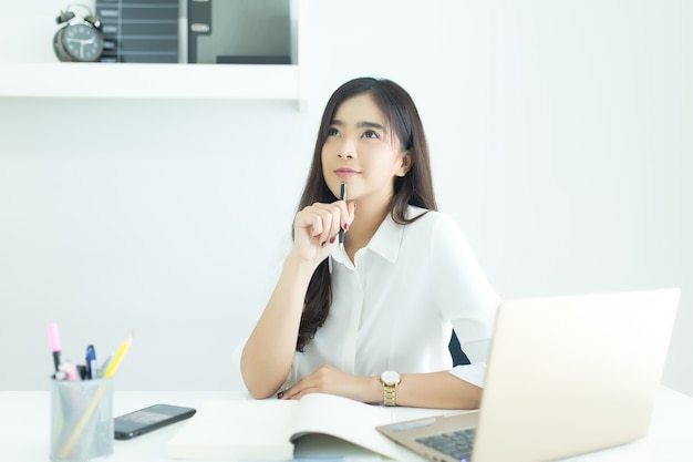 Giovane donna asiatica di affari che sorride e che pensa idea circa lavoro allo scrittorio in ufficio moderno.