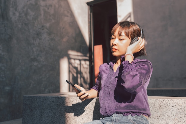 Giovane donna asiatica felice che ascolta la musica con le cuffie tramite smartphone e si diverte mentre era seduto sul lato della strada.