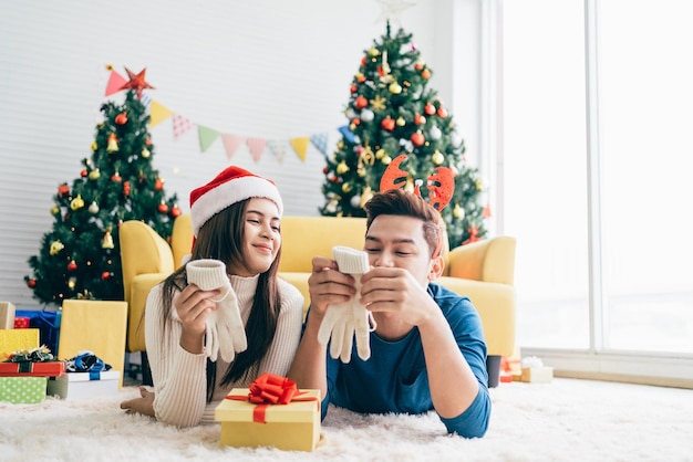 Giovane donna asiatica felice che indossa un cappello di Babbo Natale ha sorpreso il suo ragazzo con i guanti Un regalo di Natale mentre era sdraiata sul tappeto con un albero di Natale sullo sfondo