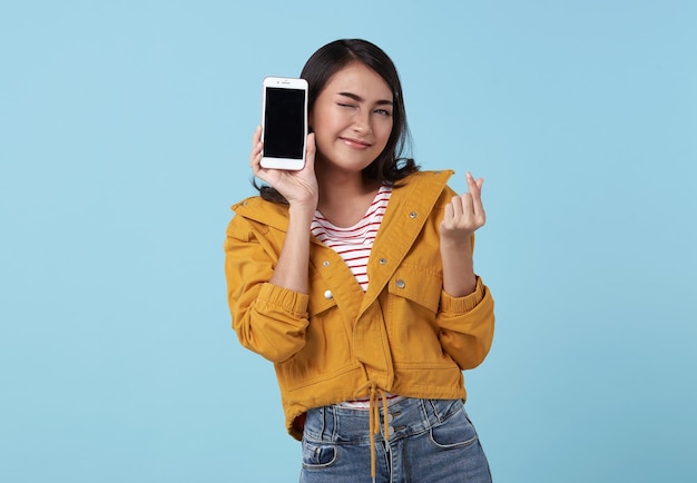 Giovane donna asiatica felice che mostra al telefono cellulare dello schermo in bianco e al mini cuore isolato sopra blu