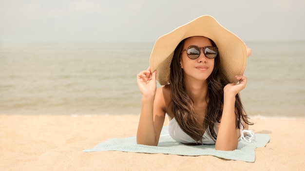 Giovane donna asiatica felice con il cappellino da sole che si trova alla spiaggia di estate