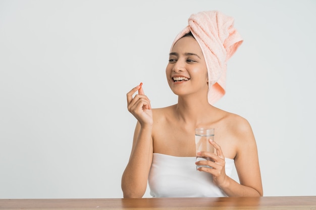 Giovane donna asiatica giovane di bellezza che mangia pillole e acqua potabile sulla parete bianca.