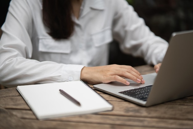 Giovane donna asiatica in abbigliamento casual elegante che scrive sul taccuino e lavora al computer portatile mentre si siede in ufficio creativo o caffè ragazza che lavora con il computer portatile sul tavolo di legno nel caffè