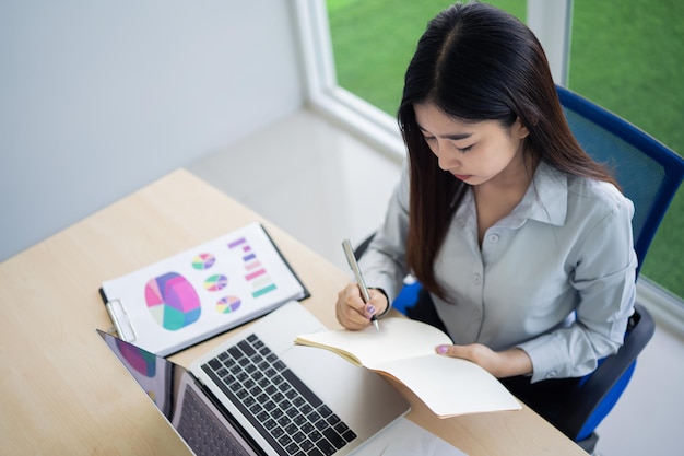 Giovane donna asiatica in abbigliamento casual elegante che scrive sul taccuino e lavora al computer portatile mentre si siede in ufficio creativo o caffè ragazza che lavora con il computer portatile sul tavolo di legno nel caffè