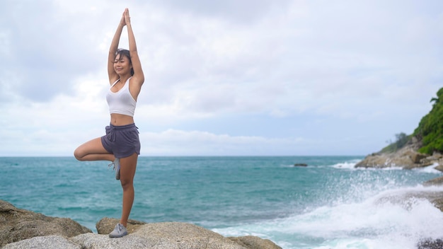 Giovane donna asiatica in abiti sportivi che fa yoga sulla roccia al concetto di salute e meditazione balneare