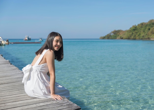 Giovane donna asiatica in abito bianco seduto sul molo di legno nel mare tropicale in giornata di sole