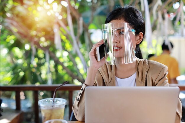 Giovane donna asiatica in abito casual con visiera per l'assistenza sanitaria, seduta in una caffetteria e utilizzando smartphone e lavorando su un computer portatile. Nuovo concetto di distanza normale e sociale