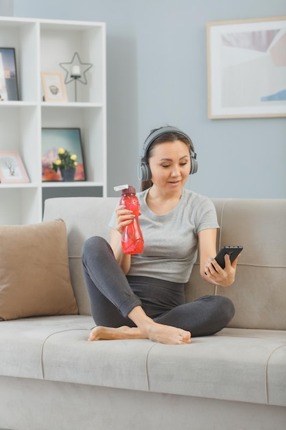 Giovane donna asiatica in buona salute con le cuffie seduta su un divano con una bottiglia d'acqua a casa stanca dopo l'allenamento utilizzando lo smartphone