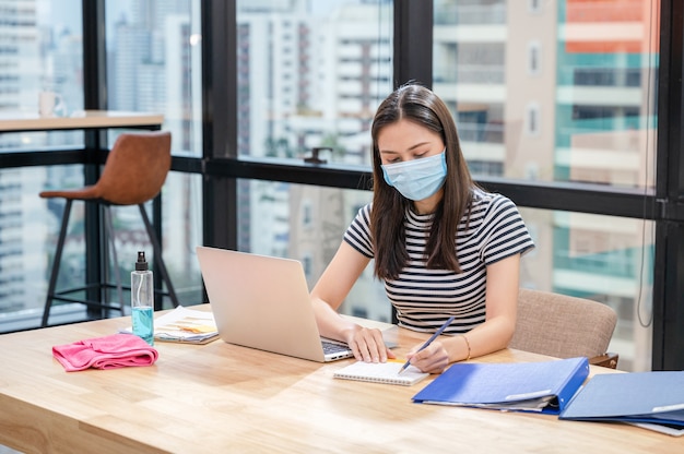 Giovane donna asiatica in casual con maschera facciale e nota di pianificazione sul calendario presso il nuovo ufficio normale