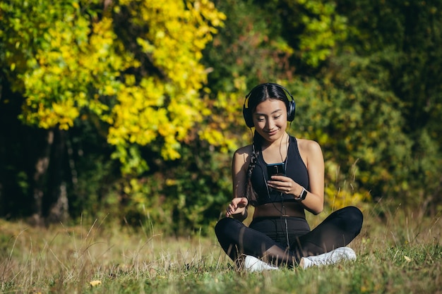 Giovane donna asiatica in forma seduta sul tappetino nella posizione del loto meditando rilassandosi e ascoltando musica all'aperto. La femmina della ragazza felice gode della natura con le cuffie nei boschi o nel parco. Guarire con i suoni