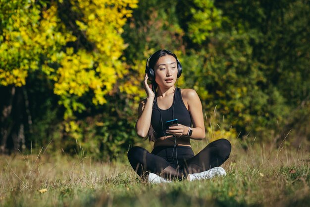 Giovane donna asiatica in forma seduta sul tappetino nella posizione del loto meditando rilassandosi e ascoltando musica all'aperto. La femmina della ragazza felice gode della natura con le cuffie nei boschi o nel parco. Guarire con i suoni