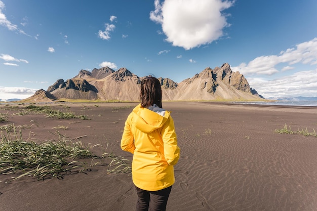 Giovane donna asiatica in giacca gialla in piedi sulla spiaggia con il monte Vestrahorn nel villaggio vichingo di Stokknes in Islanda