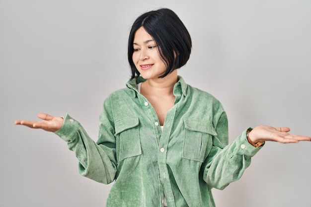 Giovane donna asiatica in piedi su sfondo bianco sorridente mostrando entrambi i palmi aperti delle mani, presentando e pubblicizzando il confronto e l'equilibrio
