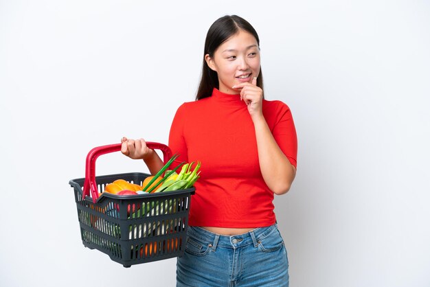 Giovane donna asiatica in possesso di un carrello pieno di cibo isolato su sfondo bianco guardando al lato e sorridente