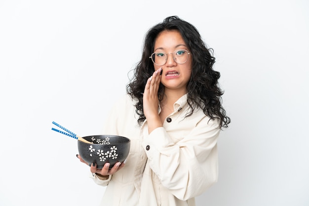 Giovane donna asiatica isolata su sfondo bianco che sussurra qualcosa mentre tiene in mano una ciotola di noodles con le bacchette