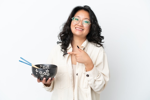 Giovane donna asiatica isolata su sfondo bianco e puntandola mentre tiene in mano una ciotola di noodles con le bacchette