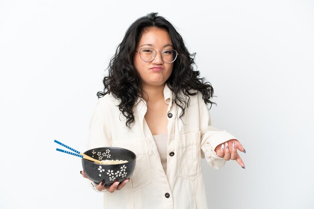 Giovane donna asiatica isolata su sfondo bianco facendo gesti di dubbi mentre si sollevano le spalle mentre si tiene una ciotola di noodles con le bacchette