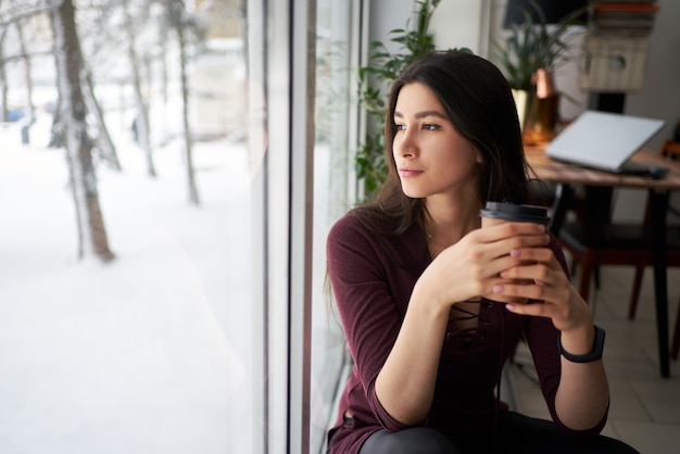 Giovane donna asiatica premurosa del brunette con la tazza di caffè che osserva attraverso la finestra in inverno