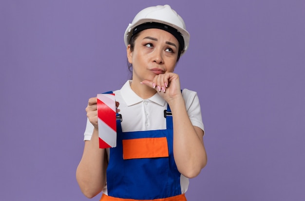 Giovane donna asiatica premurosa del costruttore con il casco di sicurezza bianco che tiene il nastro d'avvertimento e che guarda il lato