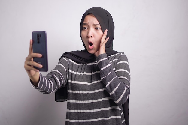 Giovane donna asiatica scioccata in maglione bianco e nero e hijab che usa il telefono cellulare e tiene la bocca con la mano isolata su sfondo bianco