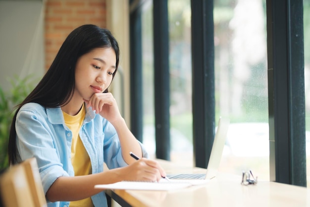 Giovane donna asiatica seduta al tavolo a studiare e scrivere sul taccuino