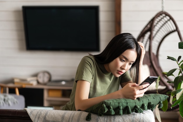 Giovane donna asiatica seduta sul divano di casa in soggiorno guardando smartphone in chat o utilizzando mo...