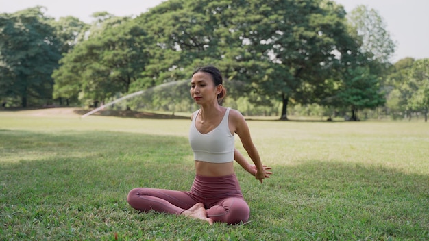 Giovane donna asiatica seduta sull'erba a praticare yoga nella posizione di posa della mucca nel parco cittadino con lo sfondo di grandi alberi Retrovisore di una donna che pratica yoga all'aperto in una giornata di sole