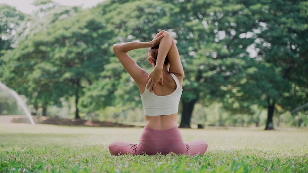 Giovane donna asiatica seduta sull'erba che pratica yoga nella posizione di posa della mucca nel parco cittadino con lo sfondo di grandi alberi. Retrovisore della donna che pratica yoga all'aperto in una giornata di sole.