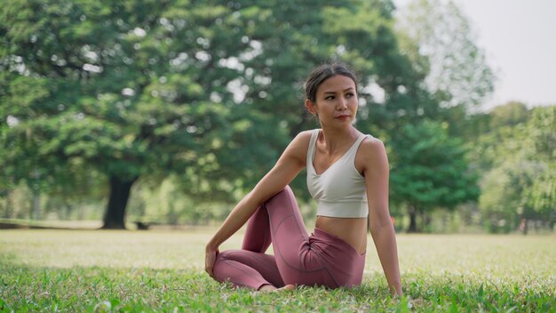 Giovane donna asiatica seduta sull'erba nella posizione del loto e alzando le mani fuori nel parco cittadino con i grandi alberi sullo sfondo. Retrovisione della pratica dello yoga femminile all'aperto in una giornata di sole.