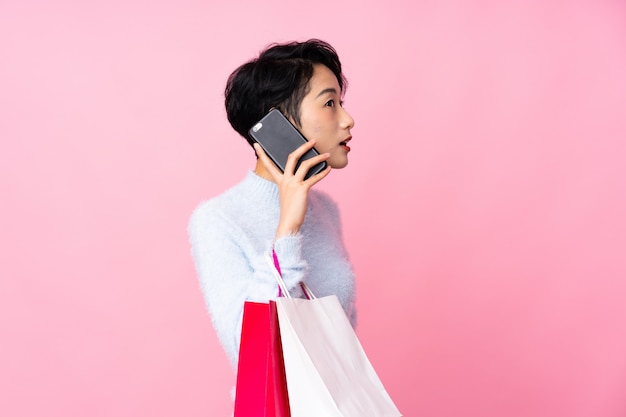 Giovane donna asiatica sopra i sacchetti della spesa rosa isolati della tenuta della parete e chiamare un amico con il suo telefono cellulare