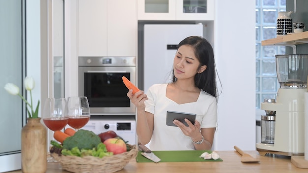 Giovane donna asiatica sorridente che legge la ricetta online sulla tavoletta digitale e prepara il pasto vegetariano in cucina
