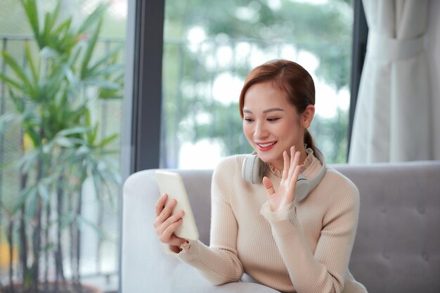 Giovane donna asiatica sorridente felice che si siede sul divano e che ha una videochiamata sullo smartphone a casa