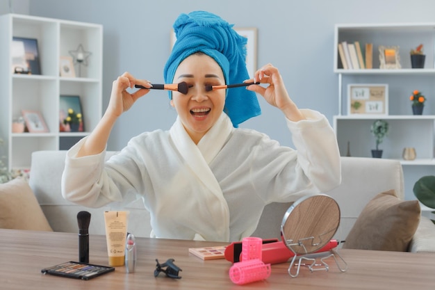 Giovane donna asiatica sorridente felice con un asciugamano in testa seduto al tavolo da toeletta a casa interni applicando ombretti o fard con specchio facendo routine di trucco mattutino