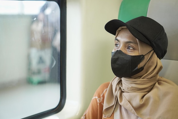Giovane donna asiatica sorridente in hijab seduto in un treno e guardando fuori dalla finestra andando in vacanza