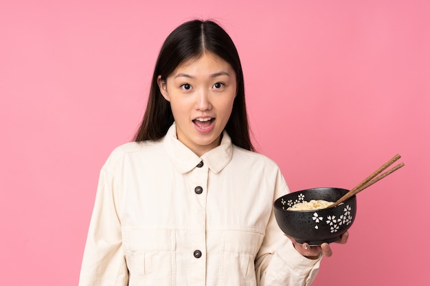 Giovane donna asiatica sulla parete rosa con espressione facciale sorpresa e scioccata mentre si tiene una ciotola di noodles con le bacchette