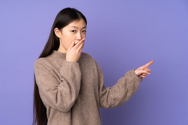 Giovane donna asiatica sulla parete viola con l'espressione sorpresa mentre indicando lato