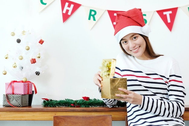 Giovane donna asiatica sveglia che porta il cappello di Santa che sorride mentre tenendo il contenitore di regalo dorato di Natale